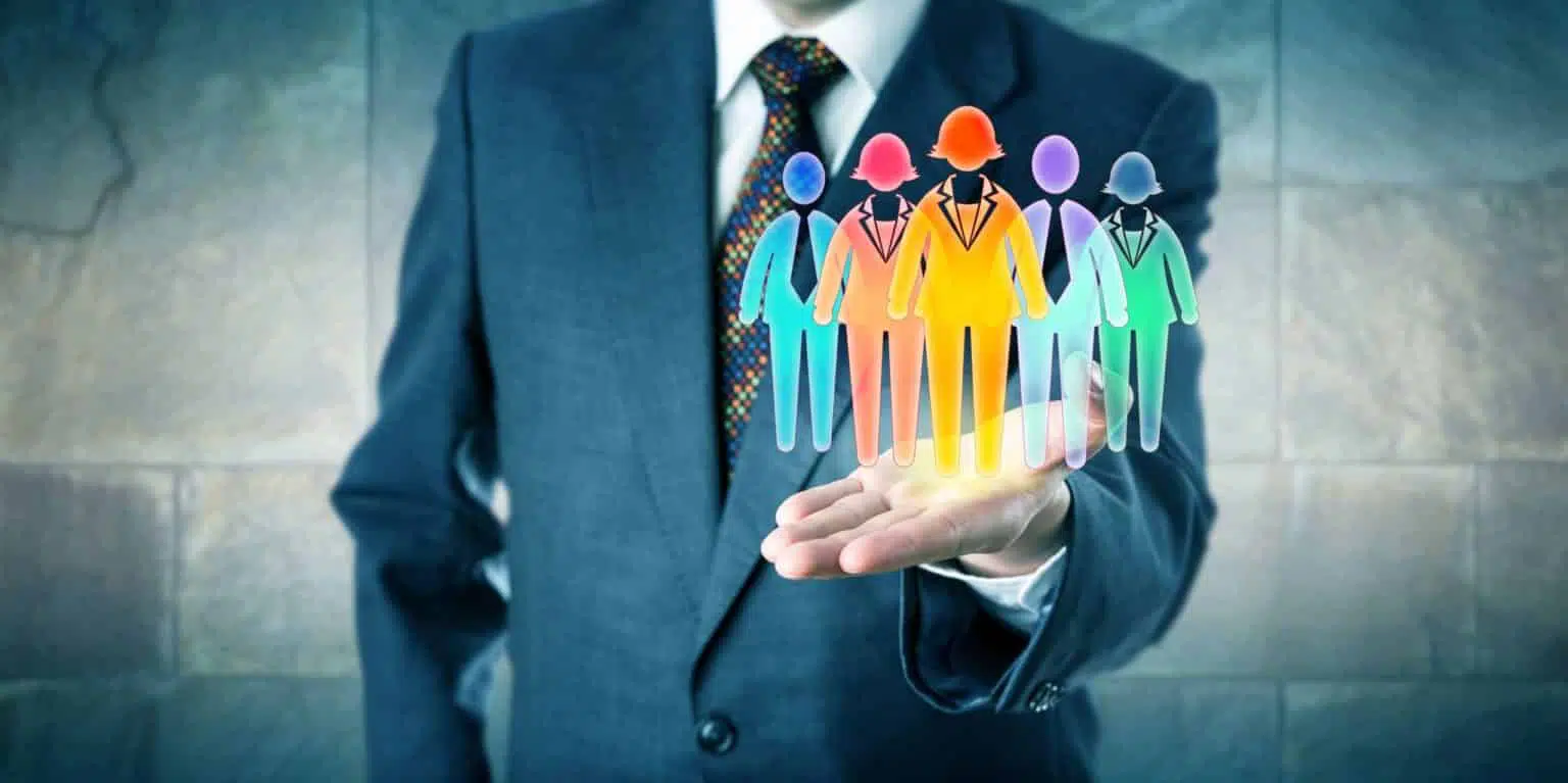 On this gray, blue, and green brick background, there is a man dressed in a grey suit with a white shirt and a black, red, yellow, and green tie with his white hand raised palm-up; he is holding a silhouette of six people in the colors yellow, red, purple, blue, and green. The image demonstrates the facility of using workforce management software to efficiently-manage the employment and deployment of customer service personnel.