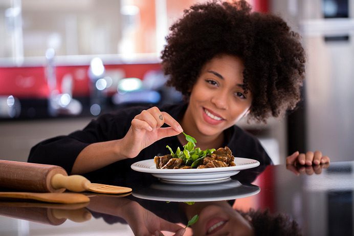 A female chef puts the final topping on an exquisite plate of filet mignon cubes with parsley while smiling to show her delight in preparing plates that guests love; with customer service outsourcing solutions, your customers within your food industry service are guaranteed to enjoy the spreading of love and joy that every chef puts as a touch on their exquisite plates.