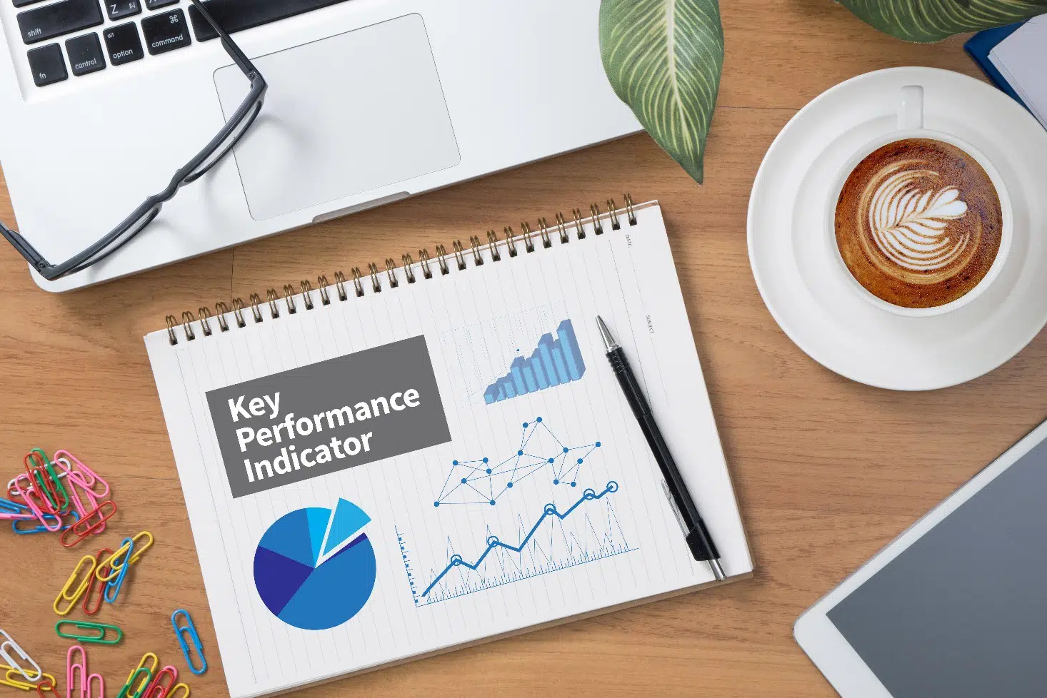 A white notepad with standard rule lines shows the words "Key Performance Indicator" which refers to key performance indicators - or, as we abbreviate it, "KPI;" the notebook contains the following: (1) a blue bar graph that shows steady growth, (2) a blue line graph that shows key growth areas, (3) a blue scatterplot graph, (4) and a blue pie graph with a slice of pie being highlighted. In addition to the detailed notebook, the picture also contains colored paperclips in the colors blue, green, pink, red, yellow, and white, a gray Apple / Mac laptop with black keys, gray glasses, a white and gray smart tablet, a white and blue planner, a black pen, a white saucer and white coffee mug with brown coffee that features a plant design prepared via cream, and also the leaves of a green plant. Everything in the picture is sitting on a wooden table.